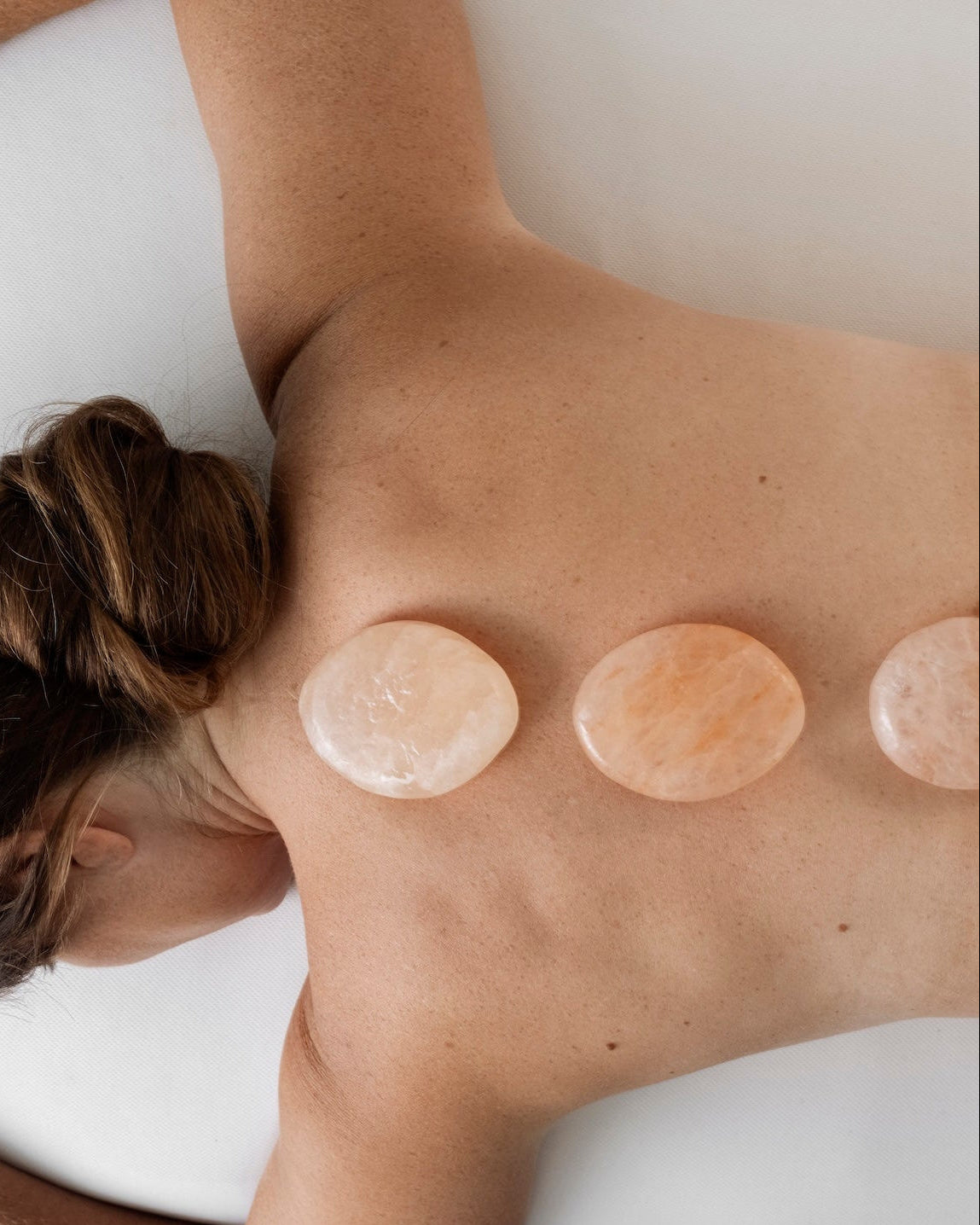 A guest at Bella Sante in Boston receiving a Himalayan stone massage while resting on a white spa table, with three smooth peach-colored stones placed along the spine.
